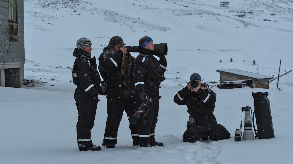 A man in a snowmobile suit takes a photo through a large telephoto lens resting on the shoulder of another man.